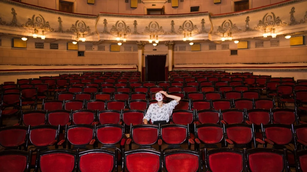 O sală de teatru veche, elegantă, cu rânduri lungi de scaune roșii goale, iar în mijloc stă o singură persoană, cu fața estompată, într-o cămașă deschisă la culoare, care pare obosită sau copleșită, cu mâna dusă la frunte.