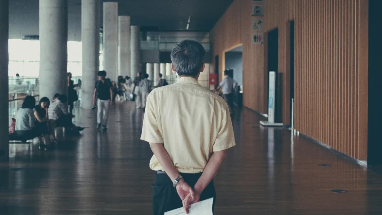 Un profesor văzut din spate, stând într-un hol spațios și modern, cu mâinile la spate ținând o foaie. În jur sunt coloane de beton, podea lucioasă din lemn și pereți cu panouri verticale din lemn. În fundal se văd mai multe persoane care circulă sau stau pe bănci,